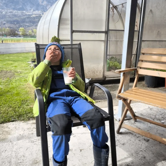 Ein Kind sitzt auf einem Stuhl in einem Gewächshaus, trinkt Milch, und in der Ferne befindet sich ein Grasfeld und ein Berg.