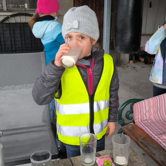 Ein junges Mädchen in einer Warnweste trinkt Milch aus einem Glas auf einem Tisch mit anderen Gläsern, in einer Außenumgebung.