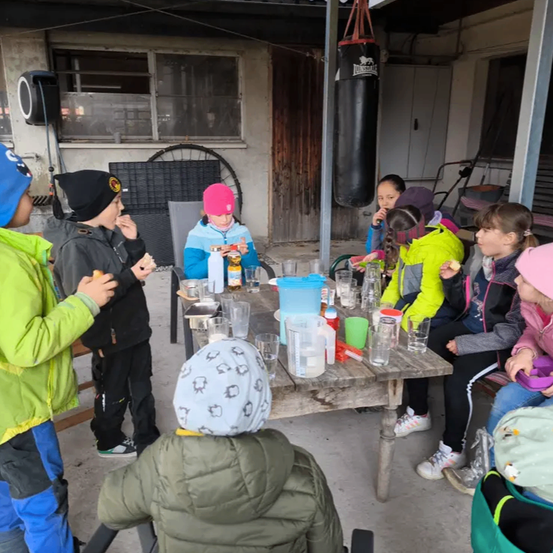 Kinder in Winterkleidung sitzen um einen Tisch im Freien, mit Getränken und Snacks. Ein Kind hält ein Sandwich. Im Hintergrund hängt ein Boxsack.