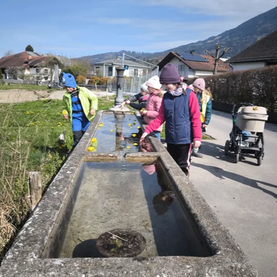 Kinder spielen mit Wasser in einem großen Betontrog, ein Kinderwagen in der Nähe. Sie tragen Winterkleidung, und Berge sind im Hintergrund.
