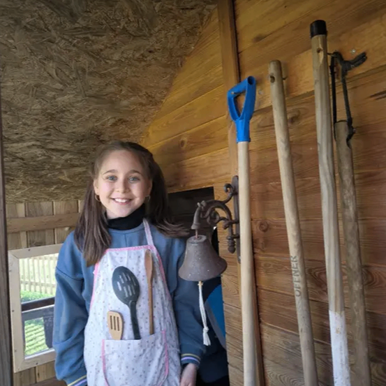 Ein junges Mädchen in einem Schürze mit Utensilien in der Tasche steht in einem Holzschuppen. Eine Glocke, eine Schaufel und ein Rechen sind an der Wand montiert.