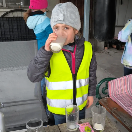 Ein junges Kind trinkt Milch aus einem Becher auf einem Tisch und trägt eine neongelbe Sicherheitsweste. Hinter ihm stehen zwei andere Kinder, eines davon trägt eine rosa Mütze. Die Szene spielt sich im Freien ab, mit einem Gebäude und einigen Möbeln im Hintergrund.