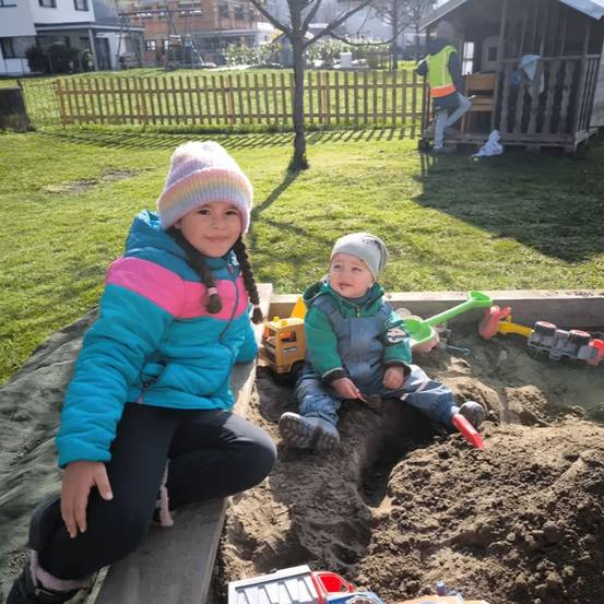 Zwei Kinder spielen in einem Sandkasten im Garten. Ein Mädchen kniet lächelnd und ein Junge sitzt mit Spielzeug in der Nähe. Ein Holzzaun und ein kleines Holzhaus sind im Hintergrund.