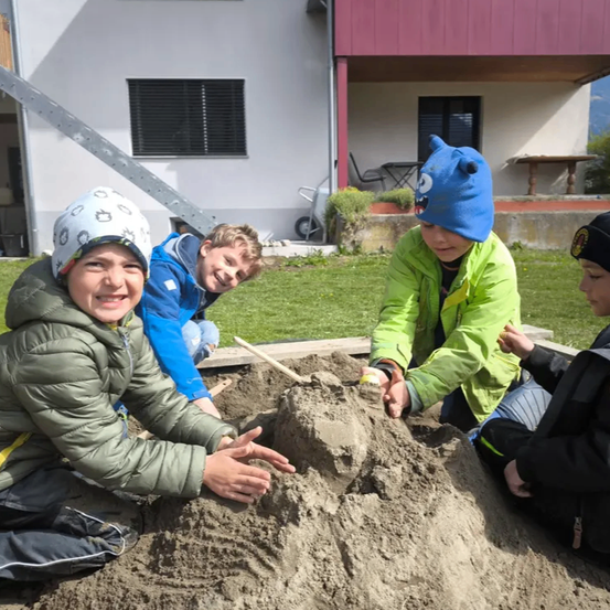 Vier Kinder spielen in einem Sandkasten im Hof. Sie tragen Winterkleidung und Mützen. Ein Kind hält eine Schaufel. Hinter ihnen steht ein Haus mit einem roten Dach.