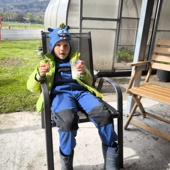 Ein Kind in einer blauen Mütze und Jacke sitzt auf einem Stuhl, hält ein Glas Milch und einen Snack in der Hand, mit einem Gewächshaus und Bergen im Hintergrund.