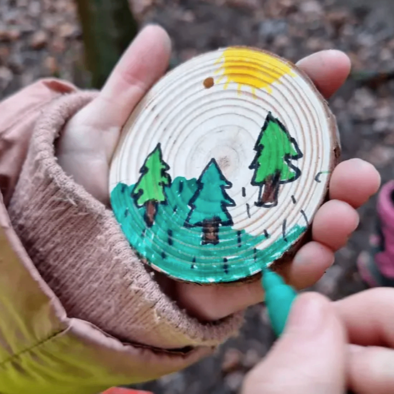 Eine Kinderhand hält eine Holzscheibe mit einer gezeichneten Landschaft. Bäume und eine Sonne sind auf der Scheibe zu sehen. Ein grüner Marker wird verwendet, um auf die Scheibe zu zeichnen.