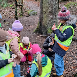 Eine Gruppe von Kindern und Erwachsenen befindet sich in einem Wald, trägt Warnwesten und Mützen. Sie scheinen zu lernen oder zu erkunden. Ein Kind hält einen roten Marker.