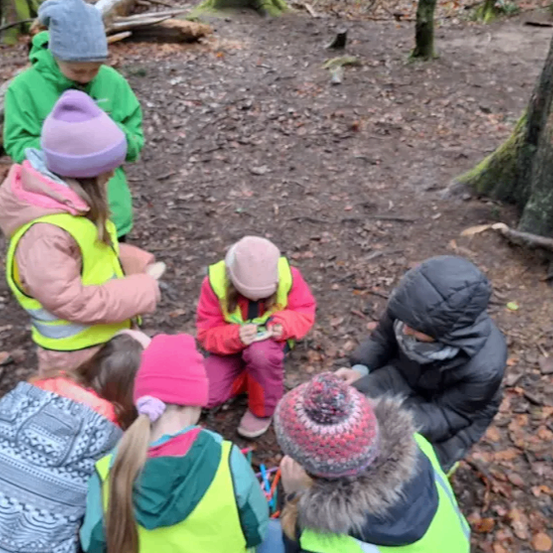 Eine Gruppe von Kindern und Erwachsenen in Winterkleidung versammelt sich im Wald, um etwas am Boden zu besprechen und zu beobachten.