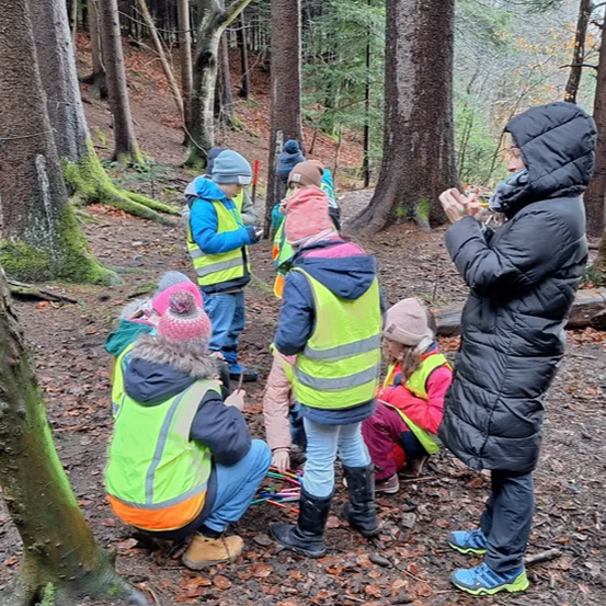 Eine Gruppe von Kindern in Warnwesten steht in einem Wald, einige halten Stöcke, und eine erwachsene Frau mit einem Stift.