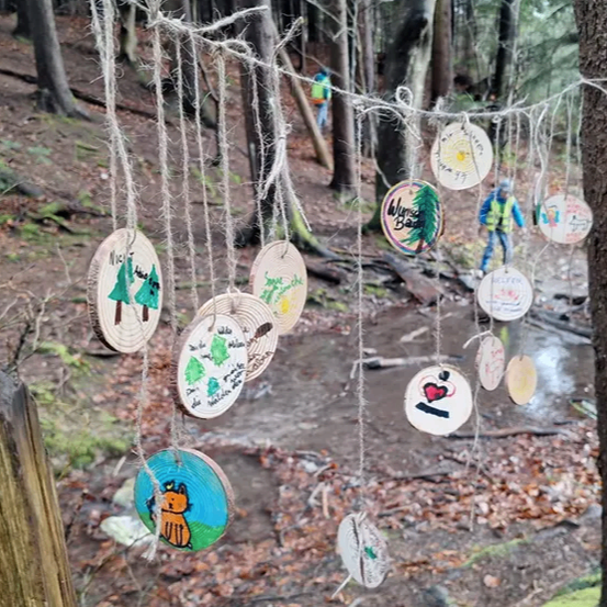 Eine Sammlung von runden Holzstücken mit verschiedenen Zeichnungen und Schriften hängt an einem Seil in einem Wald. In der Nähe geht eine Person auf einem Pfad entlang.