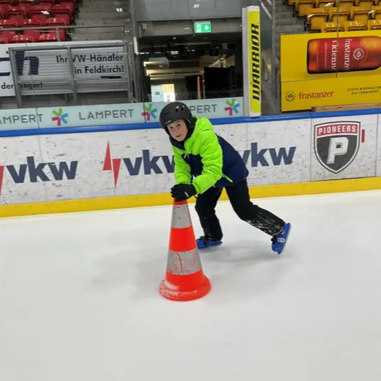 Ein Kind mit Helm übt Eislaufen in einer Arena mit Werbung an den Wänden.