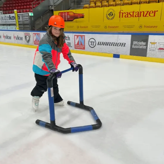 Ein junges Mädchen in einem orangefarbenen Helm übt das Eislaufen auf einer überdachten Eisbahn und hält sich an einer blau-schwarzen Stütze fest.