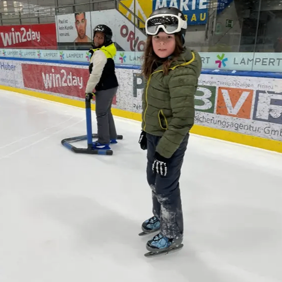 Ein junges Mädchen steht auf einer Indoor-Eisbahn, trägt eine grüne Jacke und Skibrille. Hinter ihr hält eine weitere Person einen Skilift. Die Bahn ist von Werbeanzeigen umgeben, darunter 'Win2day' und 'Marte Bau'.