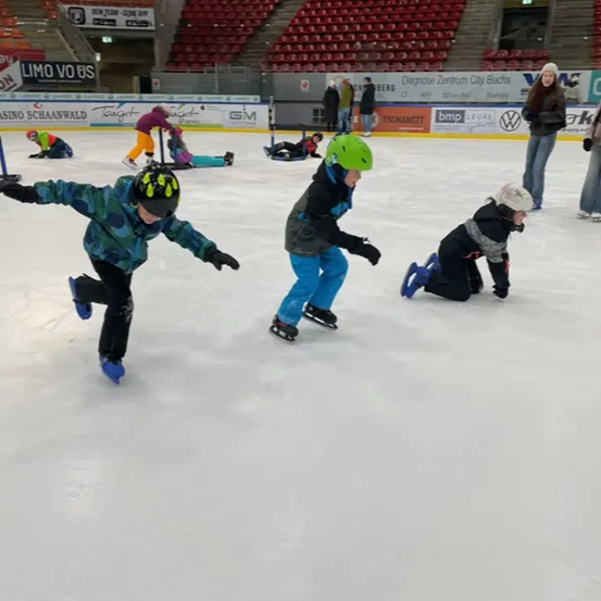 Vier Kinder üben das Eislaufen auf einer Eisbahn, tragen Helme und Handschuhe. Ein Kind liegt auf dem Eis. Im Hintergrund stehen und sitzen Menschen auf Tribünen.