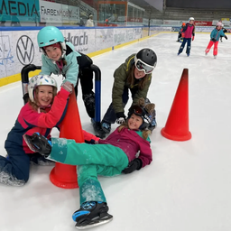 Mehrere Kinder in Helmen und Schlittschuhen üben Eislaufen in einer überdachten Eisbahn, einige liegen auf dem Eis, andere stehen in der Nähe von orangefarbenen Kegeln.