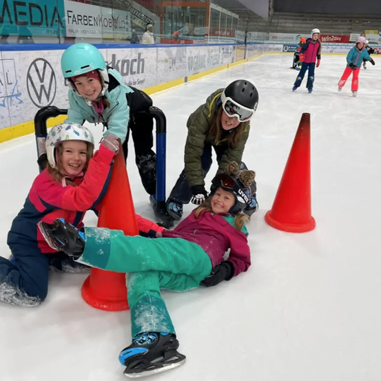 Mehrere Kinder in Helmen und Schlittschuhen üben Eislaufen in einer überdachten Eisbahn, einige liegen auf dem Eis, andere stehen in der Nähe von orangefarbenen Kegeln.