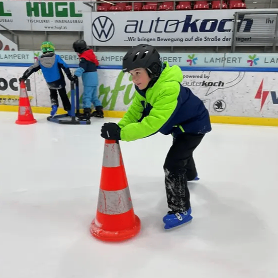Ein junger Junge in einem grün-blauen Jacken und einem schwarzen Helm fährt auf einer Eisbahn und schiebt einen orangefarbenen Verkehrskegel. Dahinter skaten auch zwei andere Kinder. Ein Werbebanner steht dahinter.