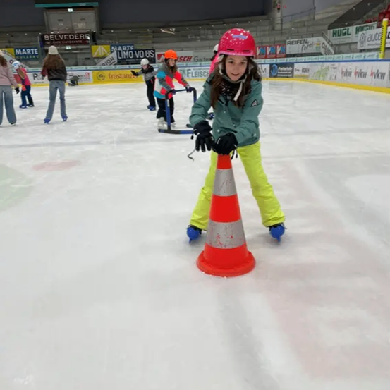 Ein Mädchen in einer grünen Jacke und gelben Hosen skatert um einen Verkehrskegel auf einer überdachten Eisbahn.
