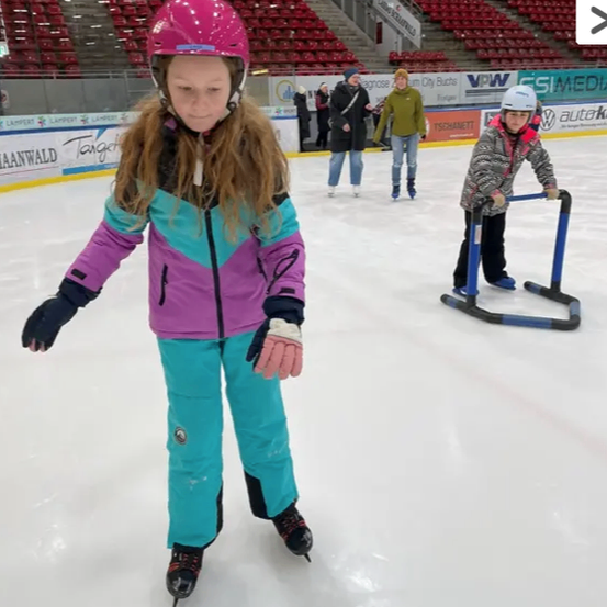 Ein Mädchen eisskat im Inneneisstadion. Sie trägt einen rosa Helm, blaue Hose und Schlittschuhe. Ein anderes Kind rutscht in der Nähe.