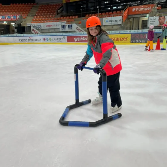 Ein Mädchen in einem roten Helm und Winterkleidung skatert auf einer Eisbahn und benutzt einen blauen und schwarzen Ständer zur Unterstützung. Die Eisbahn hat leere Tribünen und Schilder für verschiedene Sponsoren.