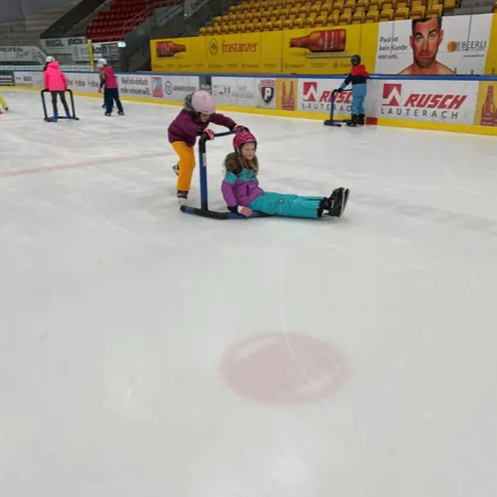 Ein junges Mädchen wird von einer Person auf einem Schlitten gezogen, während sie auf einer überdachten Eisbahn Schlittschuh läuft. Andere Menschen sind ebenfalls beim Schlittschuhlaufen. Die Eisbahn hat eine gelbe Barriere mit Werbung.