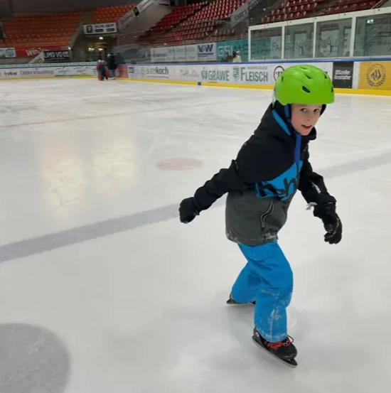 Ein junger Junge eisskatet auf einer überdachten Eisbahn und trägt einen grünen Helm, eine schwarze Jacke und blaue Hose. Die Bahn ist von leeren roten Sitzen umgeben.