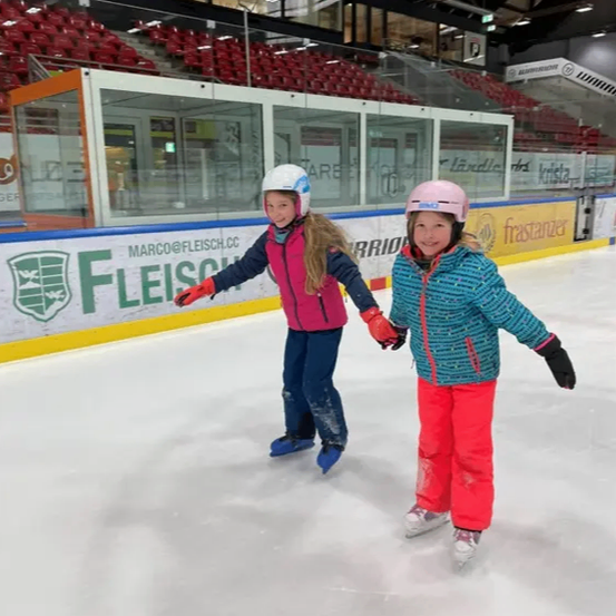 Zwei junge Mädchen sind auf einer Eisbahn beim Eislaufen. Sie halten sich an den Händen und tragen Helme und Handschuhe. Die Eisbahn hat eine blaue Barriere mit dem Wort 'Fleisch' darauf.