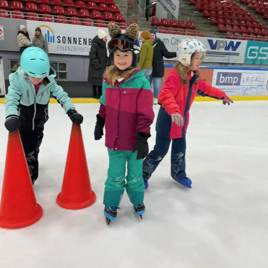 Vier Kinder rutschen in einer Eishalle auf dem Eis, umgeben von leeren roten Sitzen. Sie tragen Winterkleidung und Helme. Zwei Verkehrskegel stehen vor ihnen.
