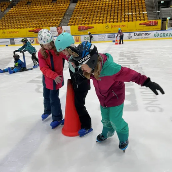 Mehrere Kinder in Winterkleidung sind auf einer Eisbahn in einer Halle Schlittschuh laufen. Sie tragen Helme und Schlittschuhe. Ein Mädchen ist auf Schlittschuhen, während andere sich an einem Kegel lehnen. Leere Tribünen umgeben die Eisbahn.