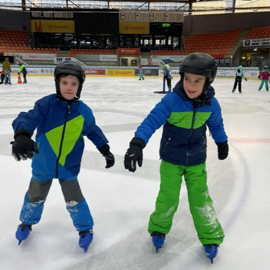 Zwei Jungen mit Helmen und Winterkleidung fahren auf einer überdachten Eisbahn Schlittschuh. Leere Tribünen umgeben die Eisbahn.