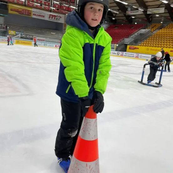 Ein junger Junge in einer grün-blauen Jacke und Helm steht auf einer Eisbahn und hält einen orangefarbenen Verkehrskegel. Hinter ihm befindet sich ein weiteres Kind auf einem Schlitten. Die Bahn hat eine gelbe Barriere, und im Hintergrund sind leere Tribünen zu sehen.