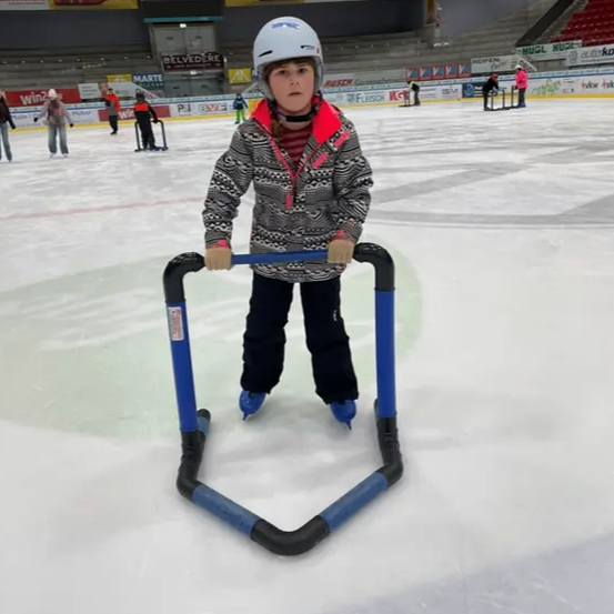 Ein Kind steht auf einer Eisbahn mit einem blauen Stützrahmen. Es trägt einen Helm und Handschuhe. Andere Skater und Zuschauer sind anwesend.