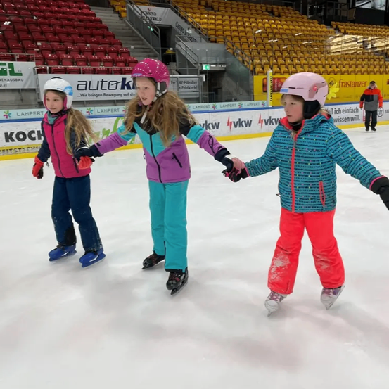 Drei junge Mädchen üben Eislaufen auf einer Indoor-Eisbahn. Sie halten sich an den Händen und tragen Helme und bunte Jacken. Im Hintergrund sind leere rote Tribünen zu sehen.