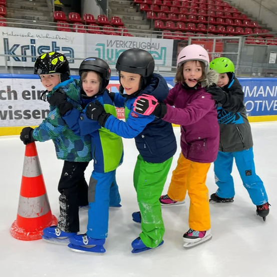 Fünf Kinder in Winterkleidung und Helmen lächeln und posieren für ein Foto auf einer Eisbahn, eines hält einen Verkehrskegel.