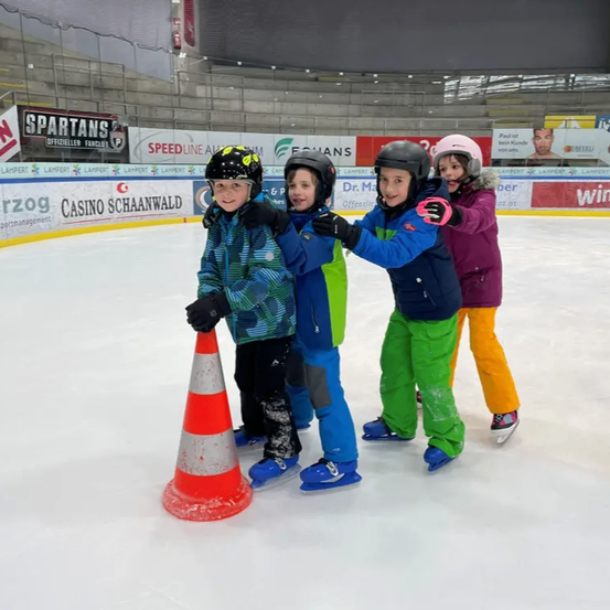 Vier Kinder in Winterkleidung stehen auf einer Eisbahn. Sie halten sich gegenseitig zum Schutz. Ein Kind trägt einen schwarzen Helm.