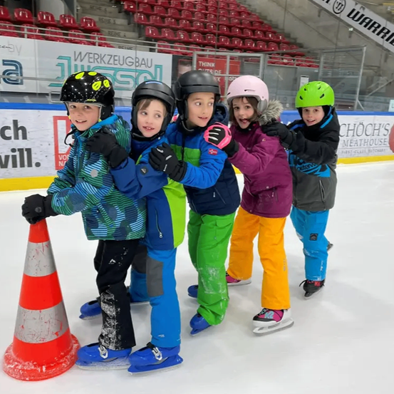 Eine Gruppe von Kindern mit Helmen und Schlittschuhen steht auf einer Eisbahn. Ein Verkehrskegel ist links. Dahinter befindet sich ein leeres Stadion.