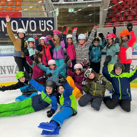Eine Gruppe von Kindern und Erwachsenen posiert für ein Foto auf einer Eisbahn, alle tragen Winterkleidung und Helme. Einige heben die Hände, während andere knien. Dahinter befinden sich leere Tribünen und ein Banner mit weißem Text.