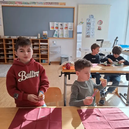 Vier Jungen sind in einem Klassenzimmer. Zwei Jungen sitzen an einem Tisch und essen. Zwei Jungen stehen, einer hält Essen.