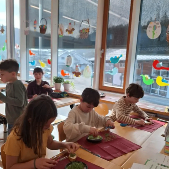 Fünf Kinder sitzen in einem Klassenzimmer um einen Tisch herum, jedes arbeitet an einem Teller mit Essen. Der Tisch ist mit rotem Tuch bedeckt, und jedes Kind hat einen Teller und Besteck. Das Klassenzimmer hat große Fenster mit farbenfrohen Dekorationen.