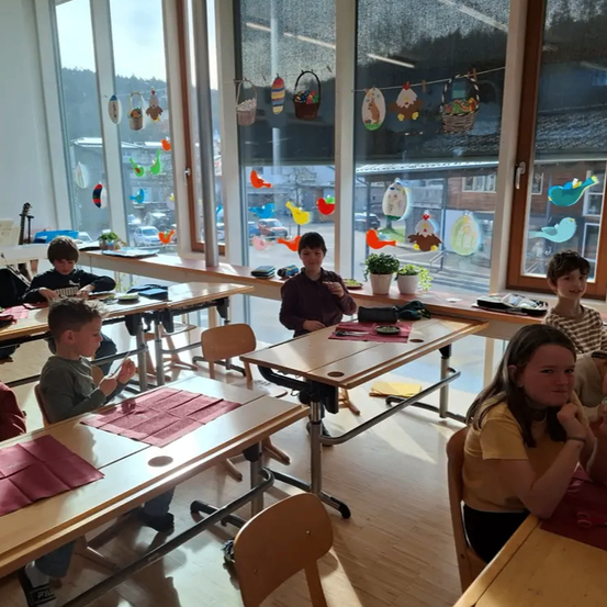 Ein Klassenzimmer mit großen Fenstern zeigt mehrere Kinder, die an Tischen sitzen. Ein Junge in der Mitte hält ein Objekt. Bunte Dekorationen hängen an den Fenstern.