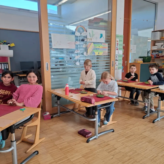 In einem Klassenzimmer sitzen mehrere Kinder an Tischen und sind in einer Aktivität verstrickt. Eine Frau sitzt auf einem Stuhl an einem Schreibtisch und schaut sich etwas an. Eine Tür ist offen und ein Regenbogen ist auf der Scheibe zu sehen.