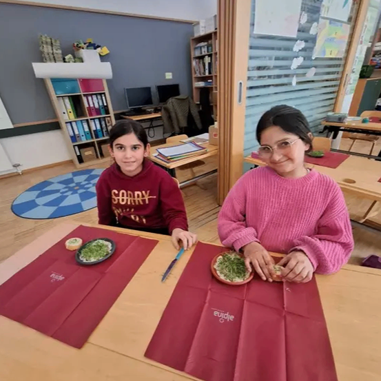 Zwei Mädchen sitzen an einem Tisch in einem Klassenzimmer. Eine trägt eine bordeauxrote Kapuzenjacke und die andere einen rosa Pullover. Sie essen Essen von Tellern mit Besteck, umgeben von roten Servietten und einer fröhlichen Atmosphäre.