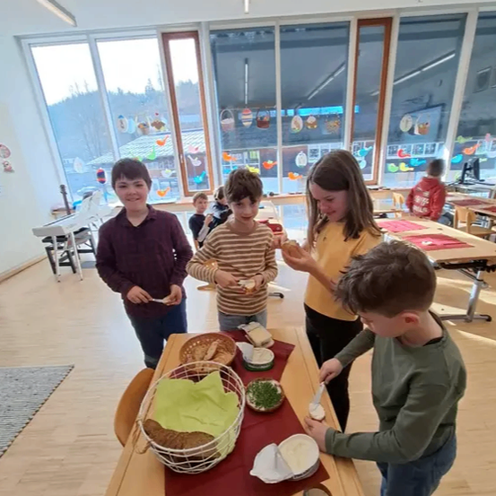 Vier Kinder versammeln sich in einem Klassenzimmer um einen Tisch, jedes hält ein Sandwich und ein Getränk. Der Tisch ist mit einem roten Tuch, einem Korb mit Brot und verschiedenen Aufstrichen gedeckt. Dahinter sind weitere Kinder zu sehen, und das Klassenzimmer ist hell mit natürlichem Licht.