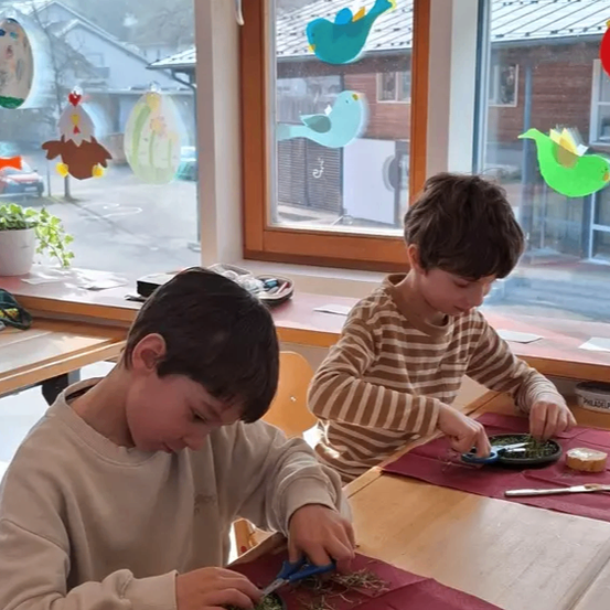Zwei Jungen sitzen an einem Tisch, einer schneidet mit einer Schere Gras auf einem Teller, der andere arrangiert Pflanzen. Sie sind in einem Klassenzimmer mit Fenstern, die mit Vogelaufklebern verziert sind.