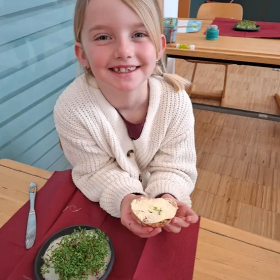 Ein junges Mädchen sitzt an einem Tisch und lächelt, während sie ein Stück Essen mit Sprossen in der Hand hält. Sie trägt einen weißen Cardigan.