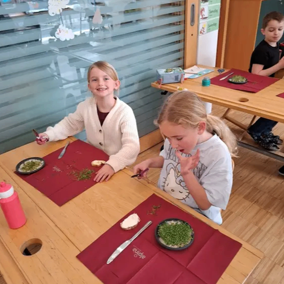 Zwei Mädchen sitzen an einem Tisch in einem Klassenzimmer, essen und benutzen Besteck. Sie lächeln und scheinen ihr Essen zu genießen. Ein weiteres Kind sitzt im Hintergrund an einem Tisch.