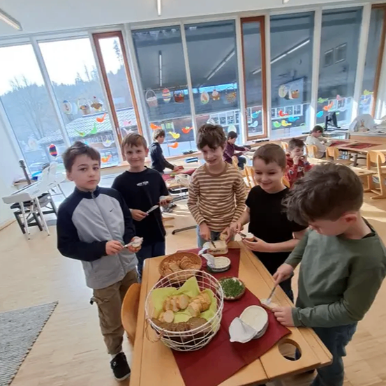 Fünf Jungen stehen um einen Tisch, jeder hält ein Sandwich. Hinter ihnen sitzen weitere Kinder an Tischen. Der Raum hat große Fenster mit Dekorationen.