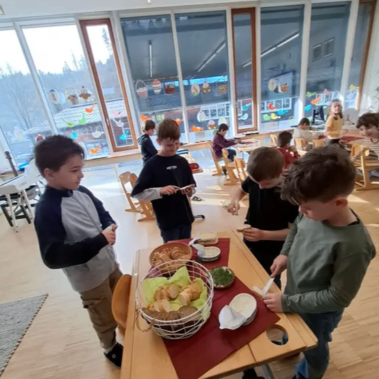 Eine Gruppe von Kindern versammelt sich um einen Tisch in einem Klassenzimmer, jeder mit einem Teller mit Essen. Der Tisch ist mit einem roten Tuch bedeckt und hat verschiedene Gerichte darauf. Sie scheinen ihre Mahlzeiten vorzubereiten oder zu genießen.