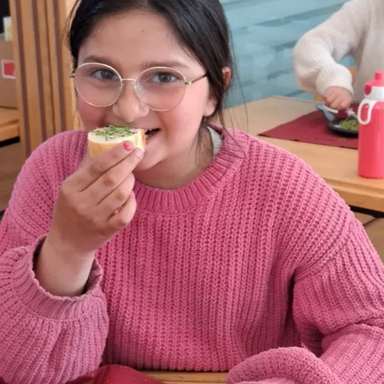 Ein junges Mädchen mit Brille isst ein Sandwich mit grüner Beilage in einem Klassenzimmer. Ein weiteres Kind ist im Hintergrund zu sehen.