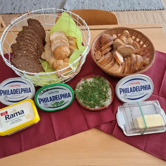 Auf einem Tisch liegt ein roter Stoff mit zwei Körben mit Brot, zwei Behältern mit Philadelphia-Frischkäse, einem Behälter mit Rama-Butter und einer Schüssel mit Petersilie.